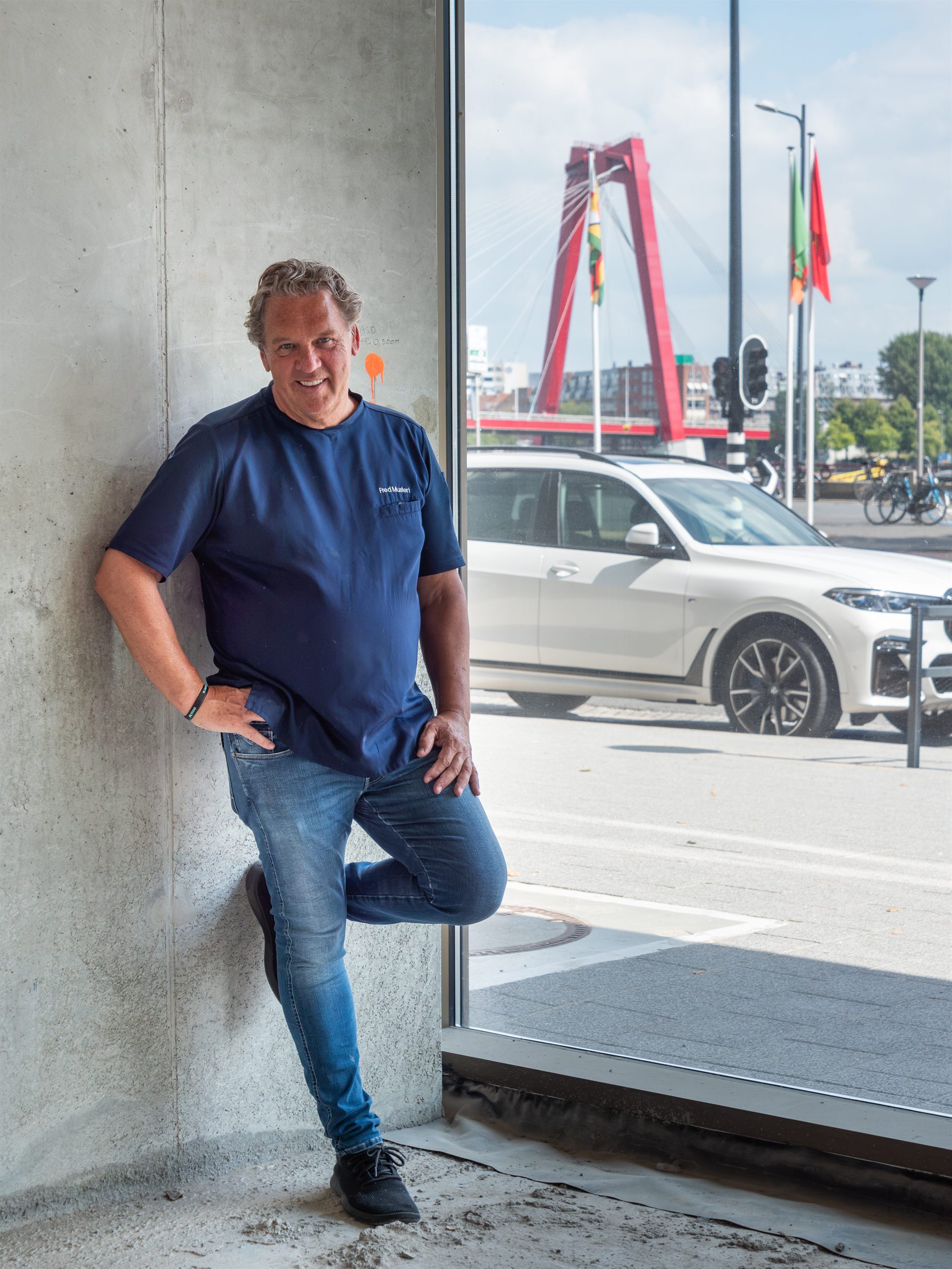 Man poseert in Restaurant Fred met de Willemsbrug zichtbaar door de glazen gevel.